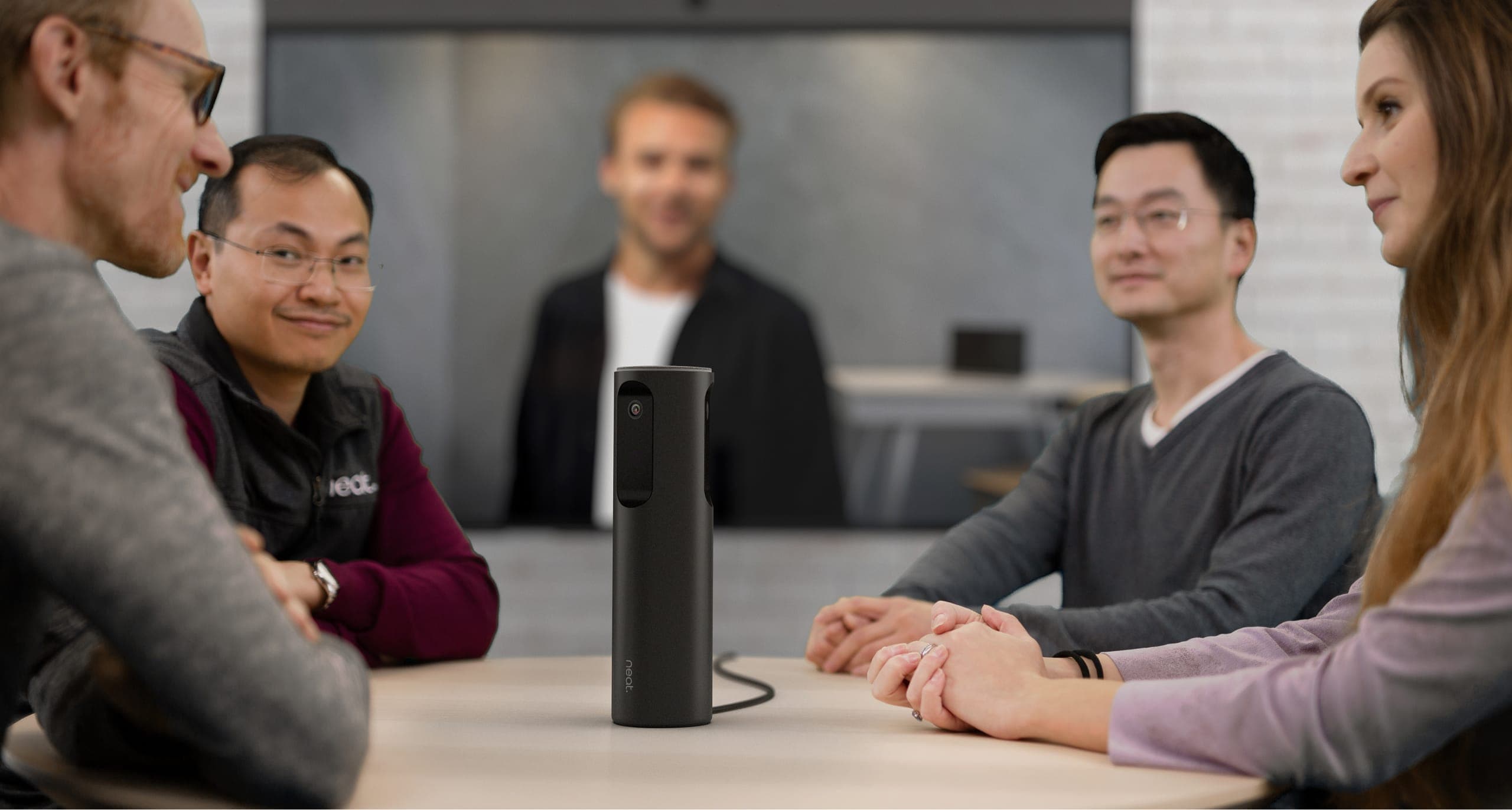Neat Center standing on a meeting-room table between four seated colleagues, with a remote participant on the wall display behind - Neat's own photograph