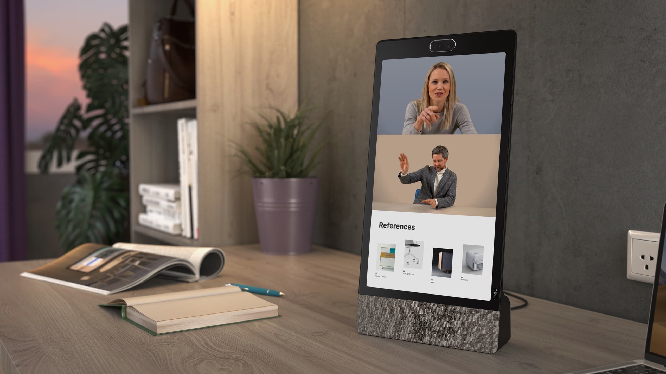 Neat Frame standing on a home-office desk beside a notebook and magazine, running a live two-person call with a shared 'References' panel on screen - Neat's own photograph; desk, plant and laptop are not included